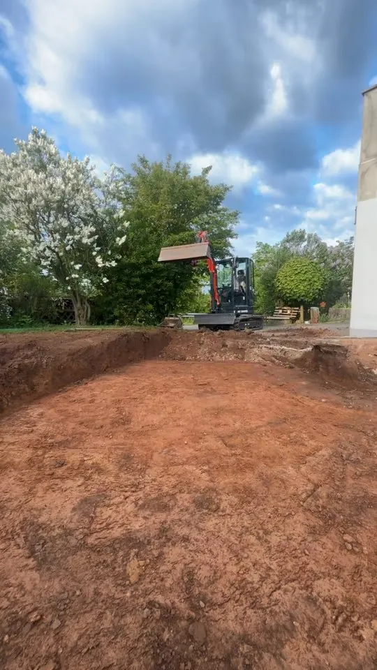Minibagger hebt eine Baugrube im Garten aus, der ausgehobene Boden ist rotbraun
