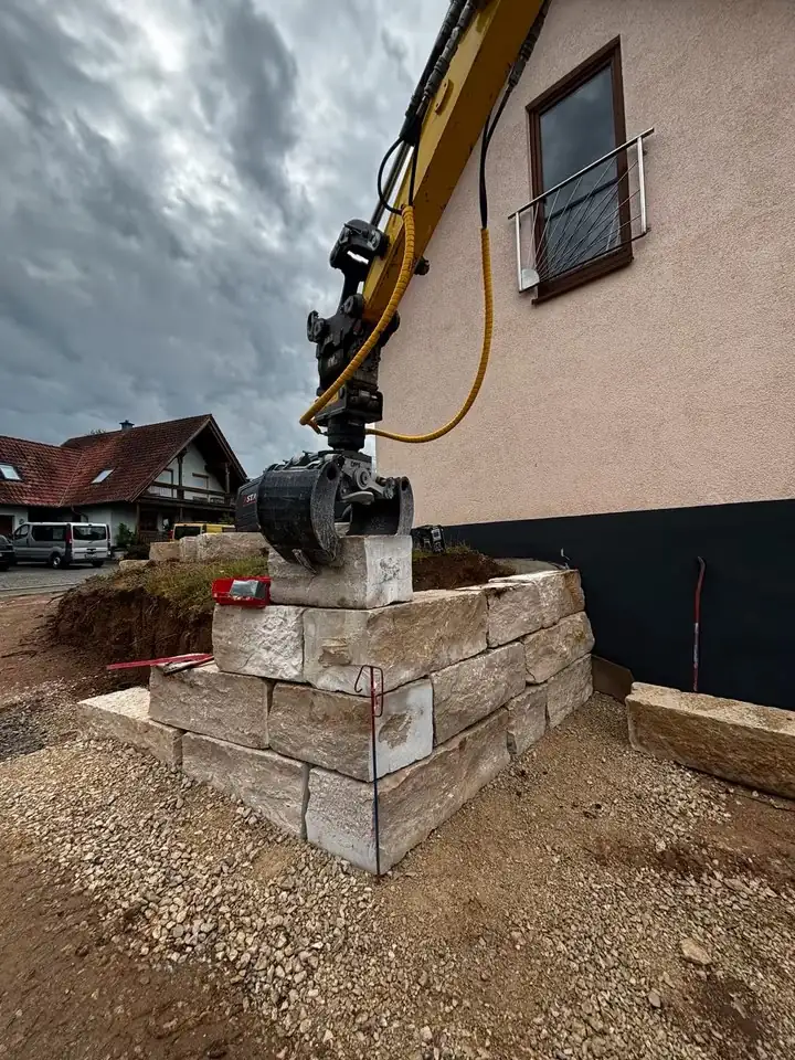 Minibagger setzt mit dem Sortiergreifer einen Steinquader auf eine Mauer