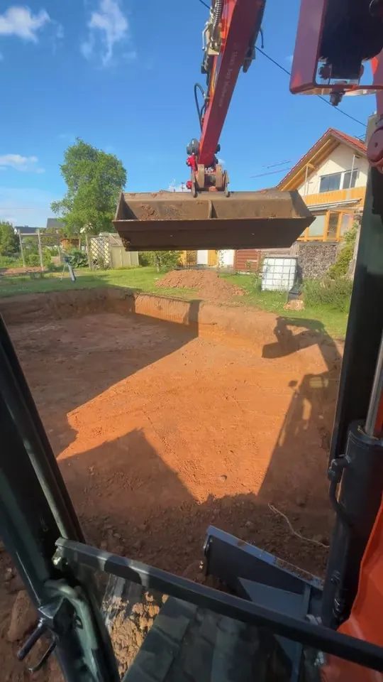 Blick aus der Baggerkabine auf eine rechteckig ausgehobene Grube im Garten