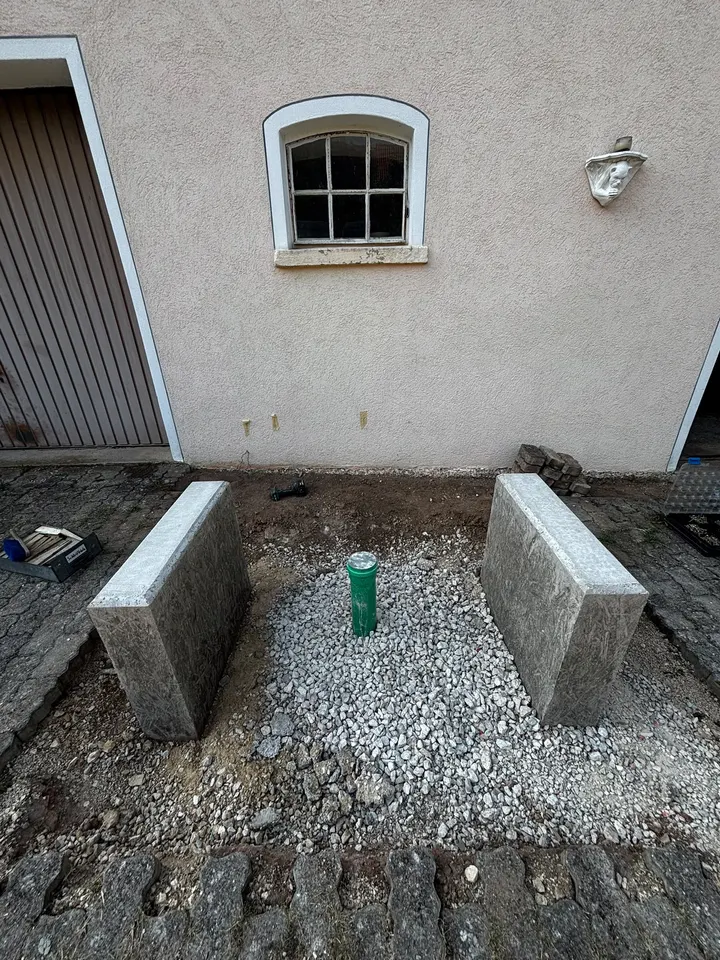 Zwei fertige Betonsockel für eine Wärmepumpe auf Kiesbett vor einem weißen Haus